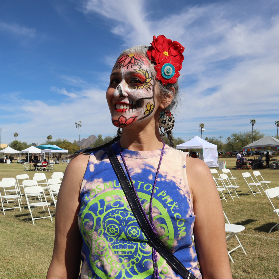Image of woman standing outside with blue sky and white clouds above and green grass and white chairs in the background. Woman has half her face painted as a calaca with red flowers in her hair and a tie-die shirt that has a dia de los muertos design on it