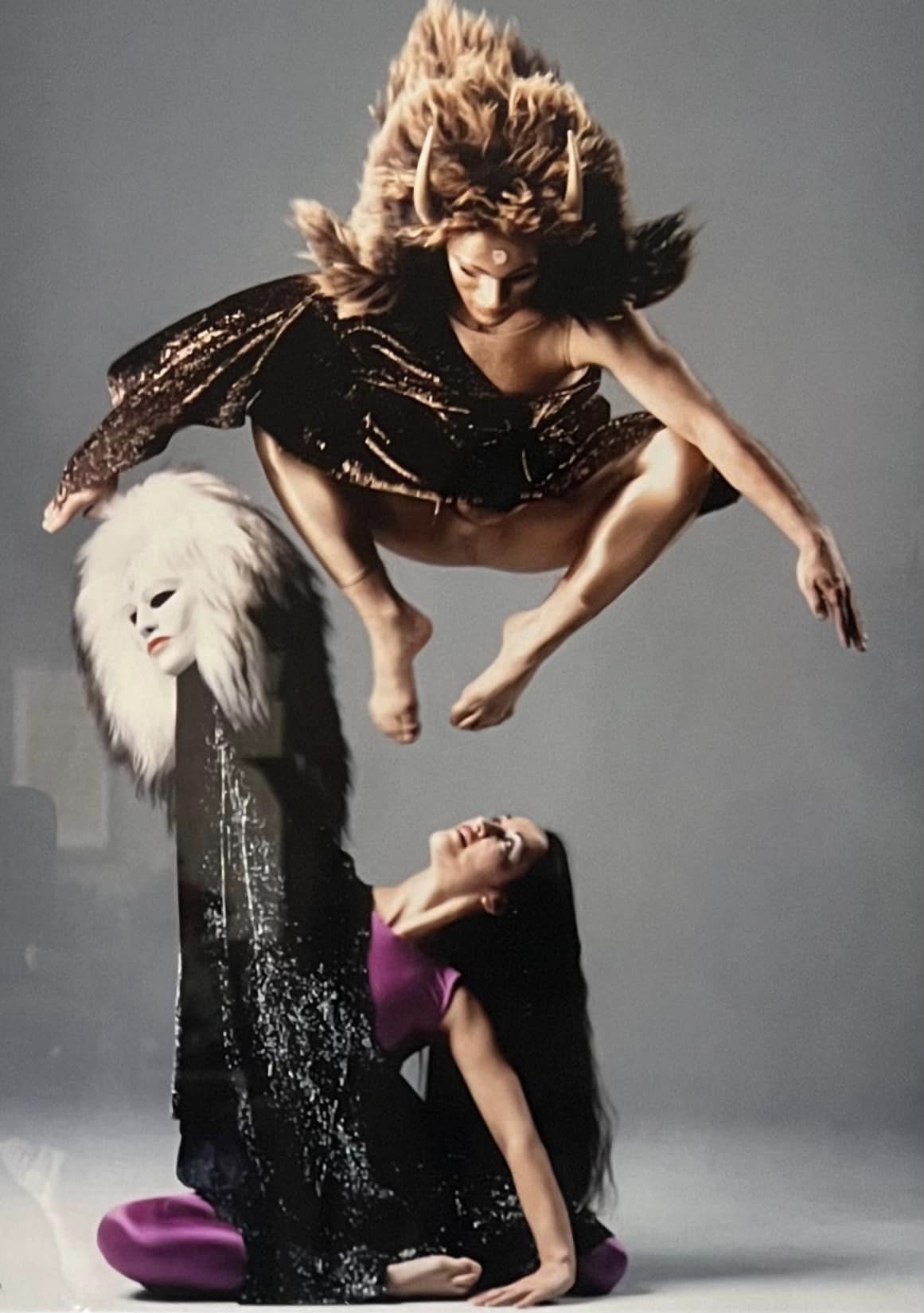 Two dancers. Mark in a fur mask in a squat position jumping in the air above Diane, sitting on the ground holding up a mask with fur