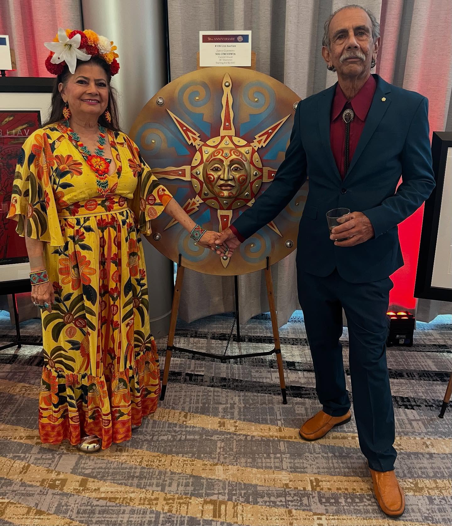 Carmen & Zarco Guerrero holding hands in front of a gold and blue circular sun sculpture by Zarco